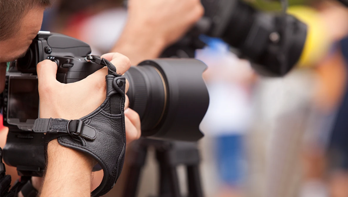 Photographer holding DSLR camera with telephoto lens and wrist strap during outdoor assignment.