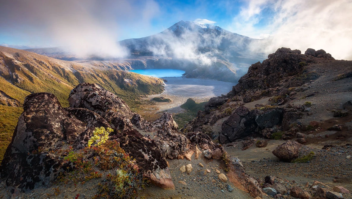 Snow-capped volcano framed by dark lava rocks and golden grassland valleys under dramatic cloudy sky.