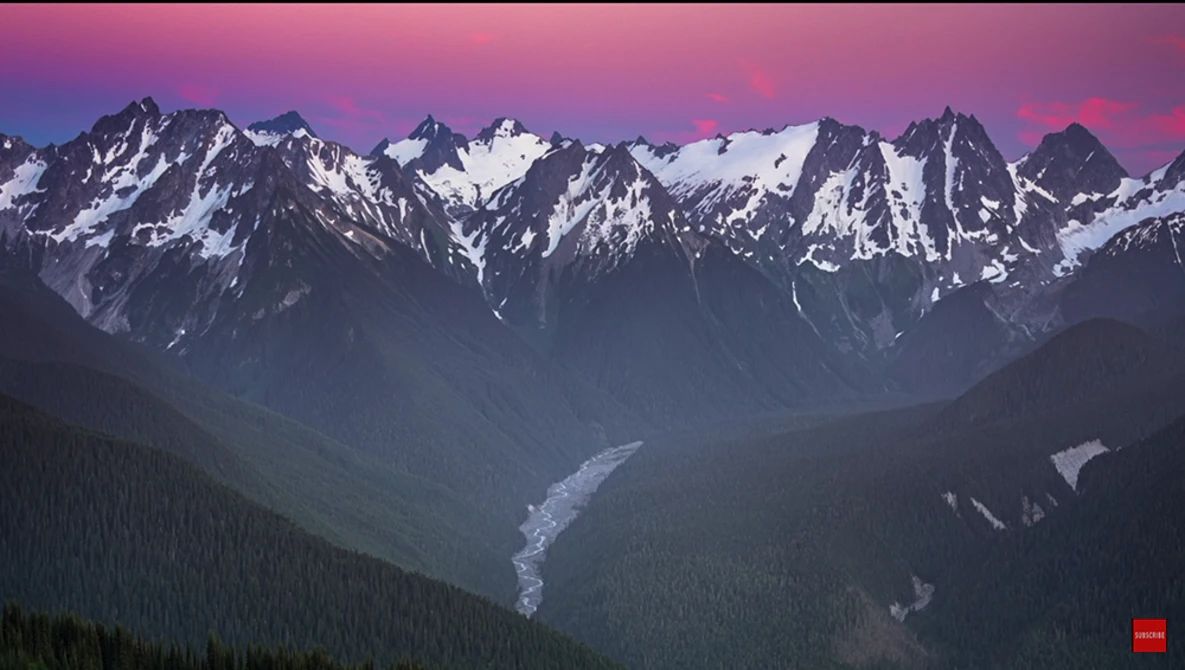 Snow-capped mountain range at dusk with a river valley and forested slopes below.