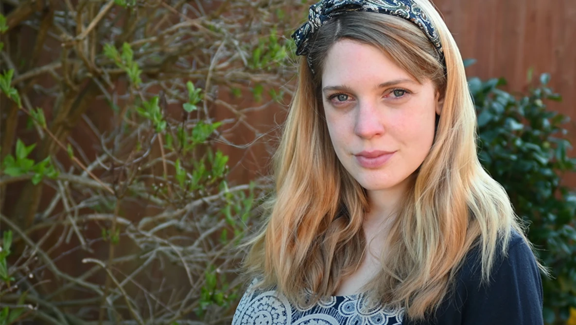 Portrait of a young woman with long blonde hair and a decorative headband against a brick and foliage background.