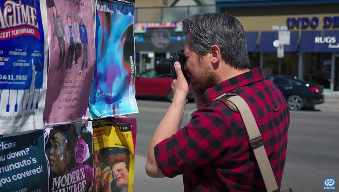 Photographer in red plaid shirt holding camera to face, composing a shot of colorful event posters on an urban street.