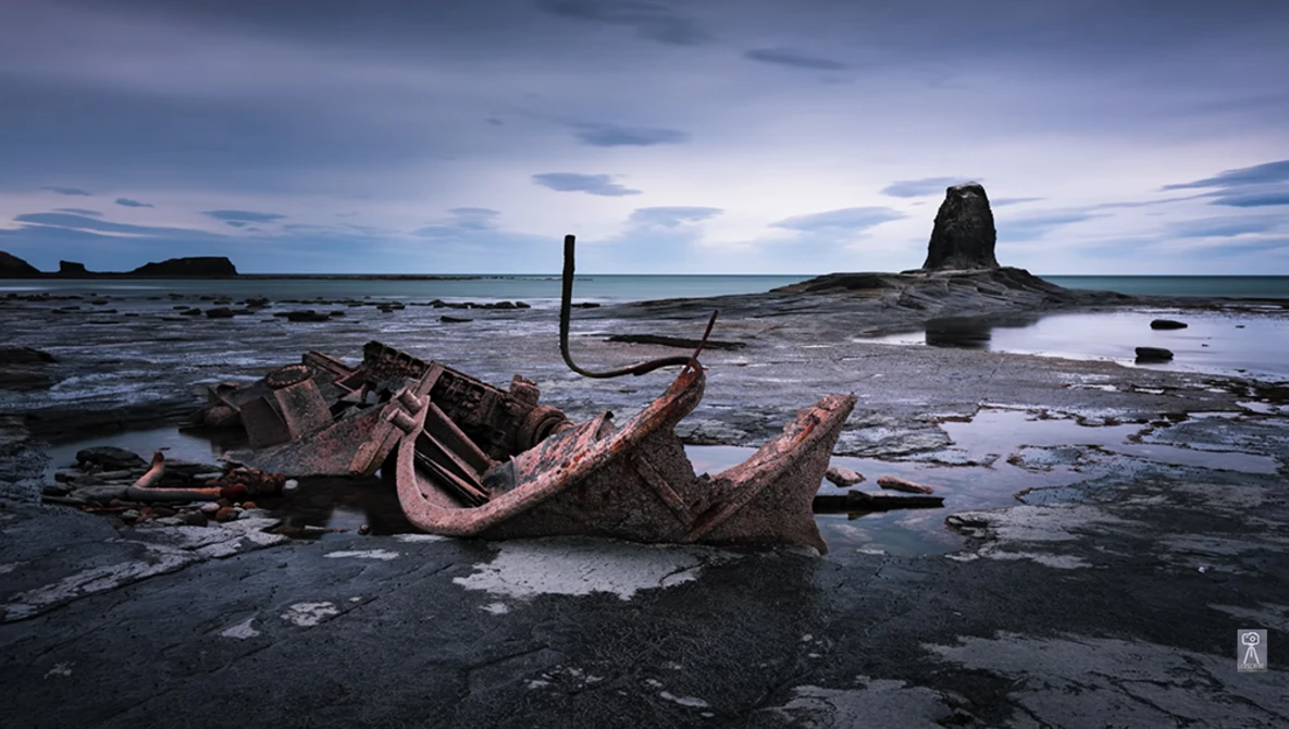 Rusted shipwreck remains on a rocky shore at twilight with a sea stack in the background.