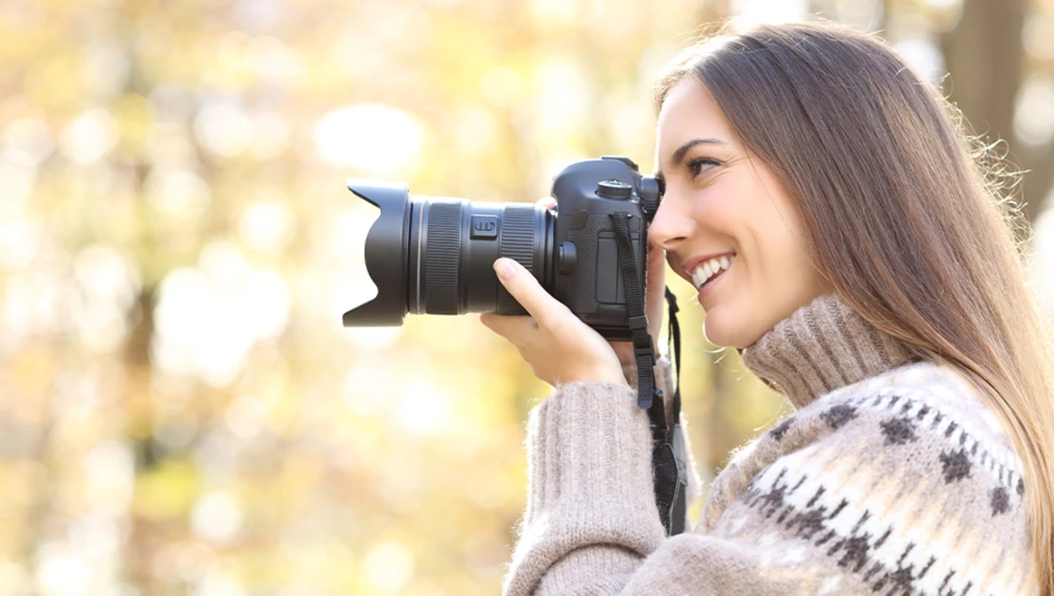 Female photographer holding a DSLR camera to her face, smiling while composing a shot outdoors.