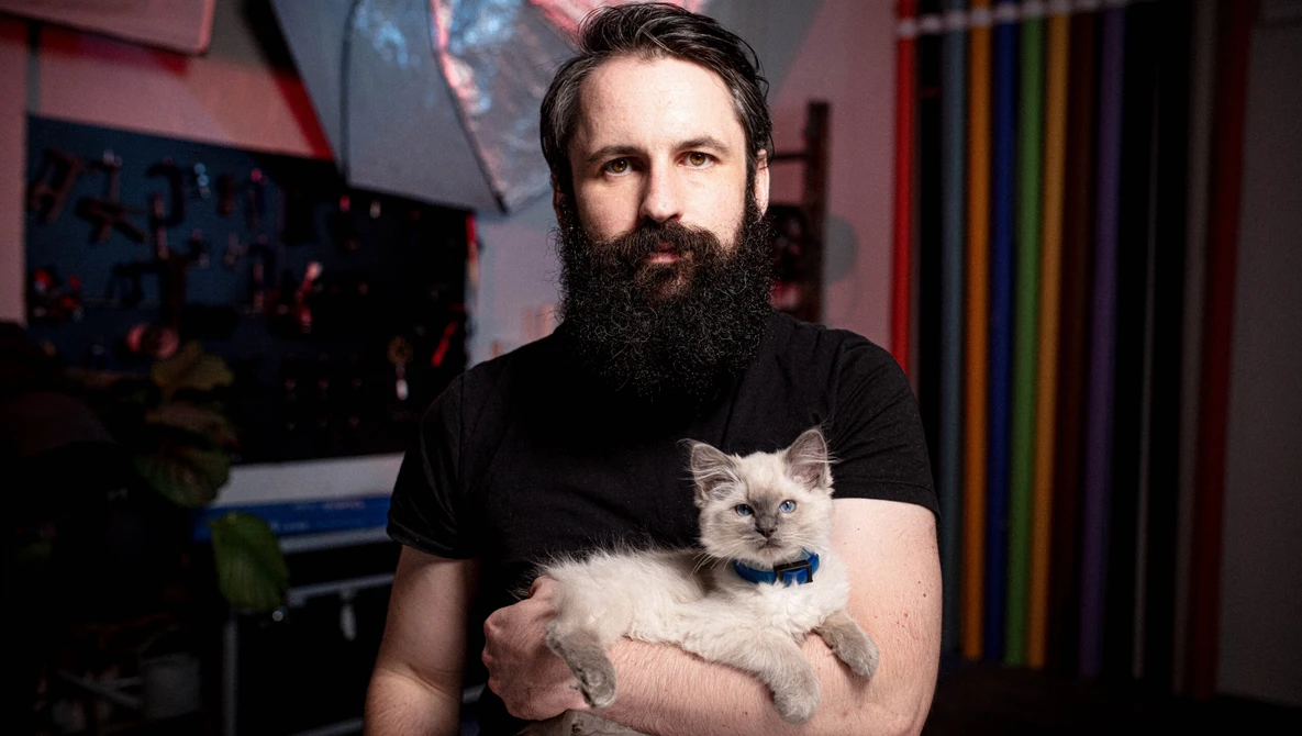 Bearded man in black t-shirt holding a light-colored kitten with blue collar in studio setting.