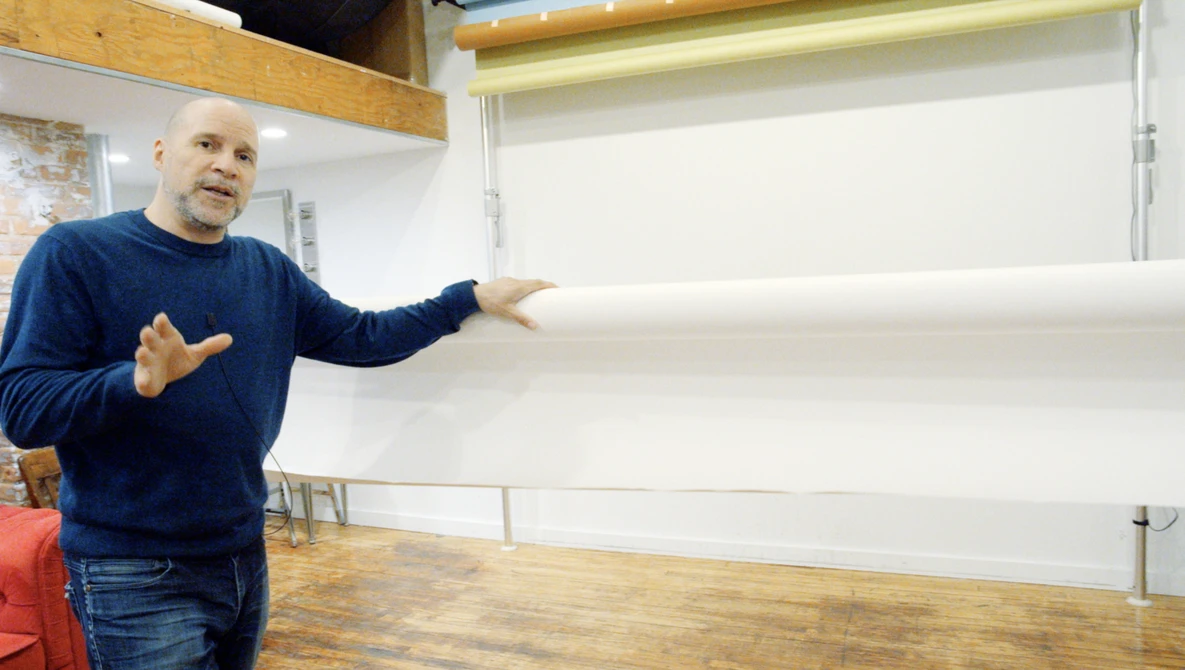 Man in navy sweatshirt gesturing toward blank whiteboard in bright studio space.