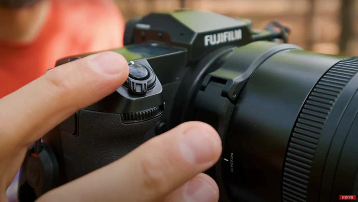 Hands holding a Fujifilm camera with telephoto lens attached, showing the shutter button and mode dial in focus.