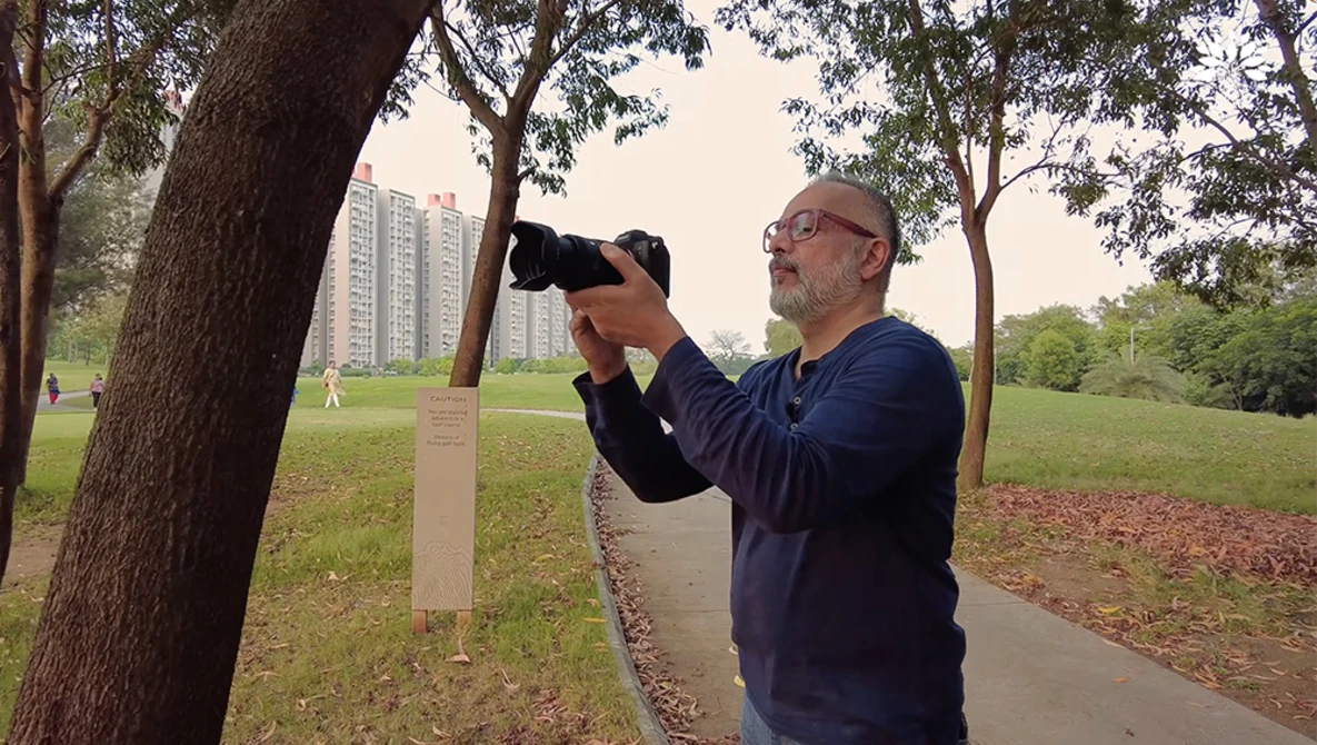 Photographer with glasses holding a telephoto lens, shooting upward beneath large trees in an urban park.