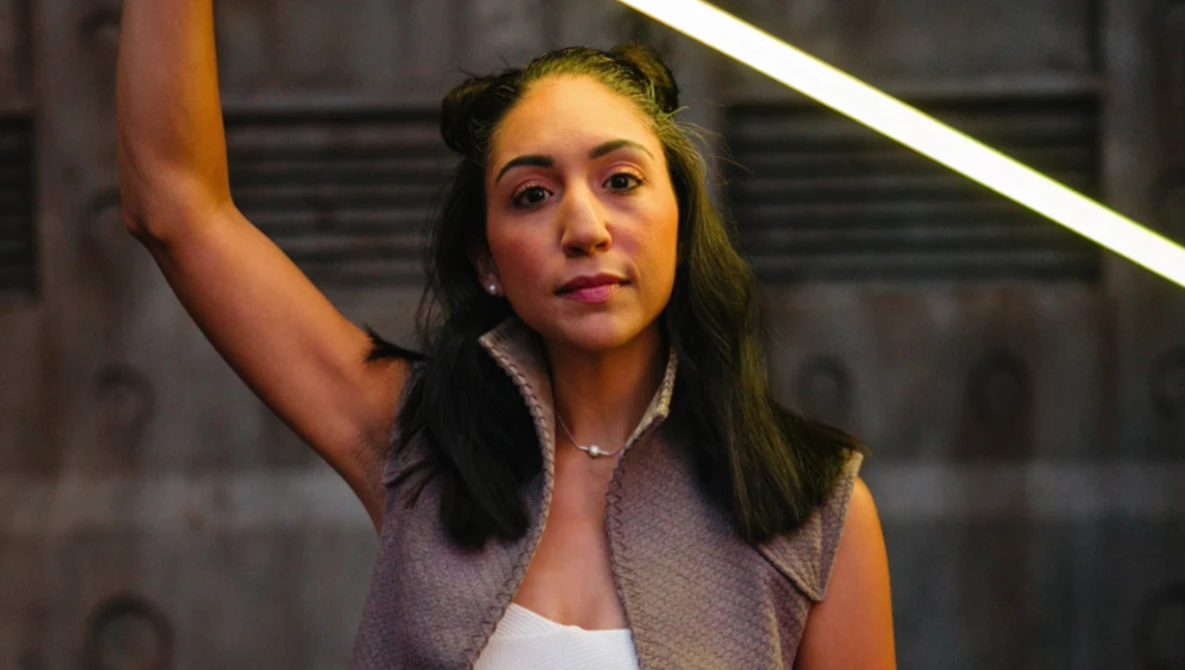 Young woman with long dark hair posing under warm neon lighting against an urban backdrop.