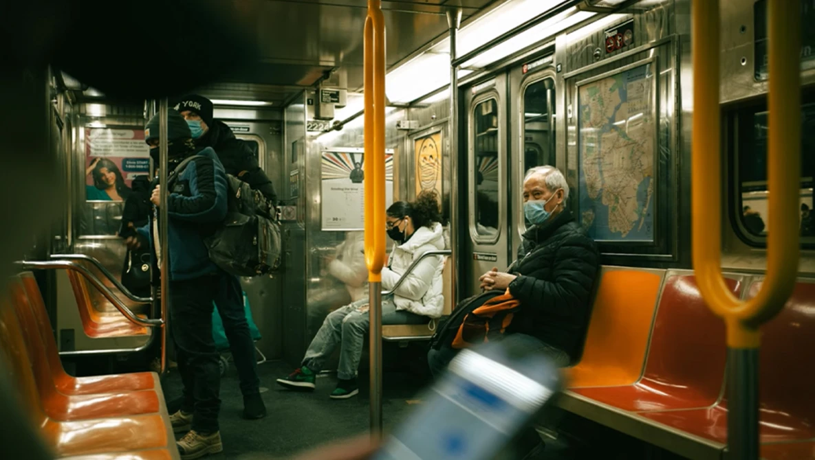 Passengers seated and standing inside a subway car with orange and green seating during daytime hours.