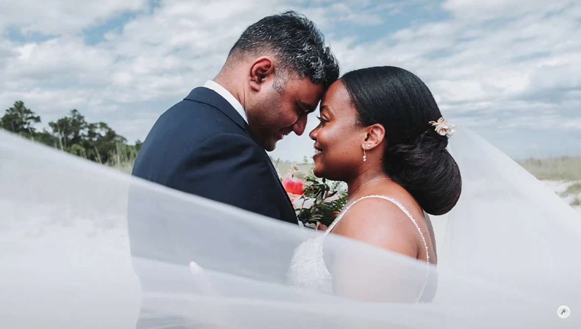 Bride and groom sharing an intimate moment outdoors, foreheads touching, with flowing veil in foreground.