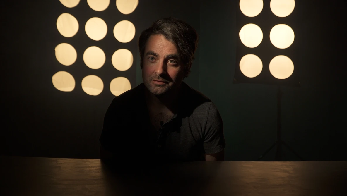 Man seated at wooden table against dark background with warm studio lighting panels.