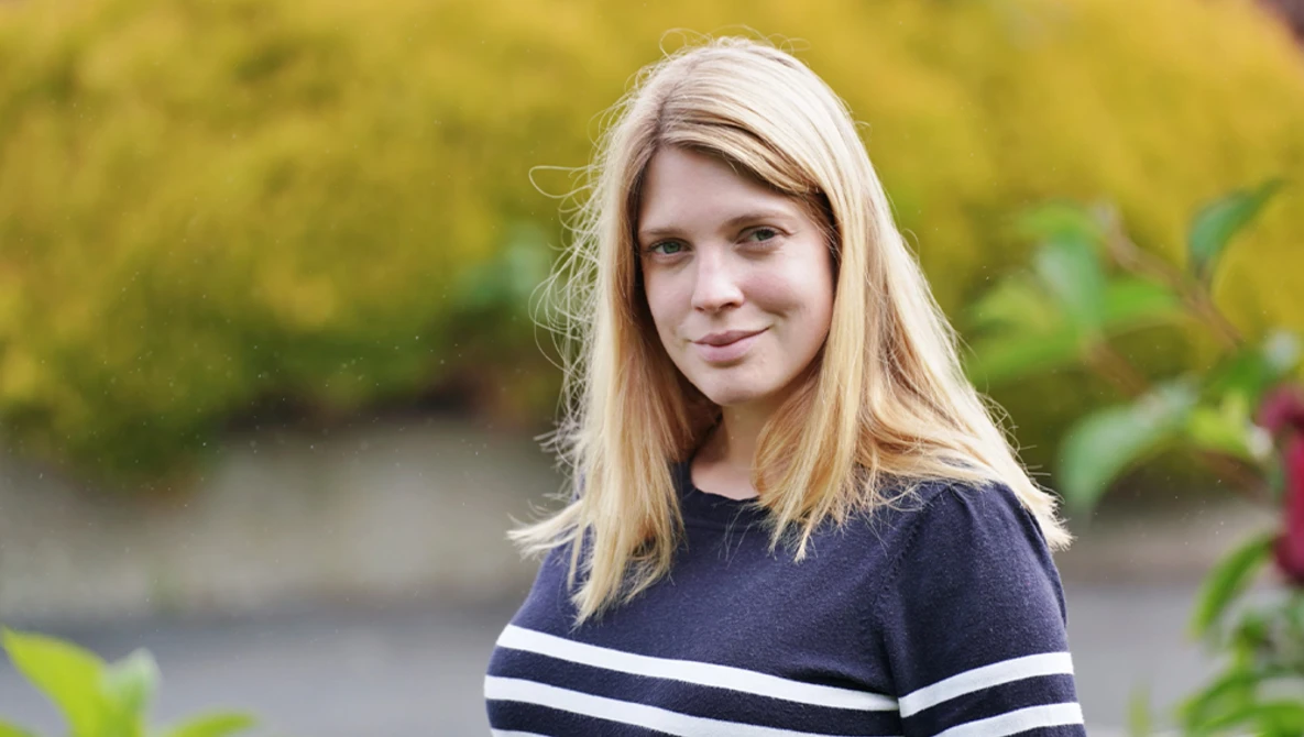 Young woman with long blonde hair wearing a dark striped sweater, photographed outdoors with soft natural lighting.