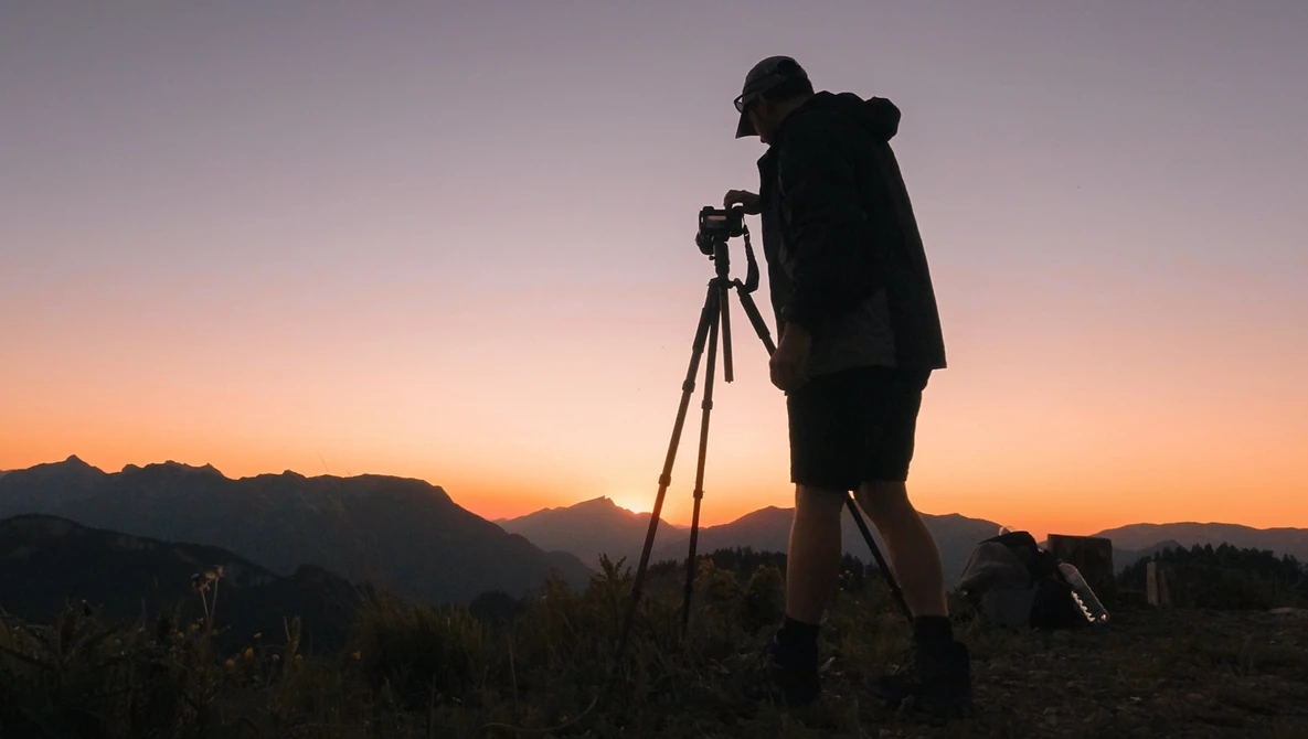 Silhouetted photographer on tripod against dramatic golden sunset sky with mountain range in background.