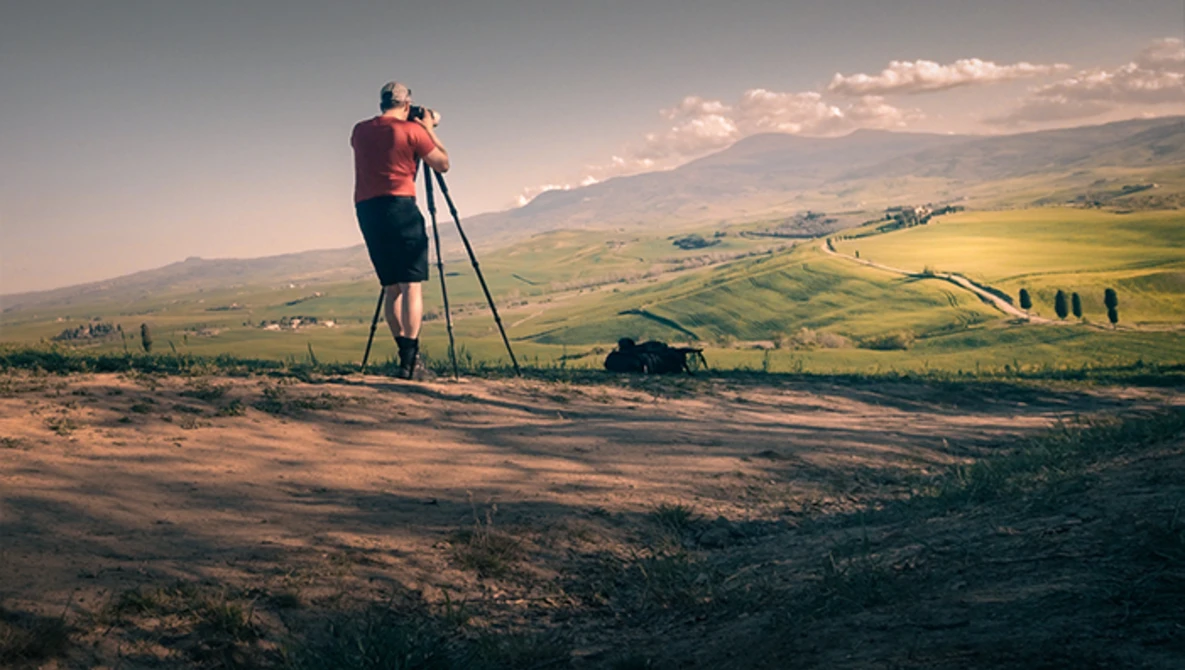 Photographer on tripod capturing landscape from elevated vantage point overlooking rolling hills and valley.