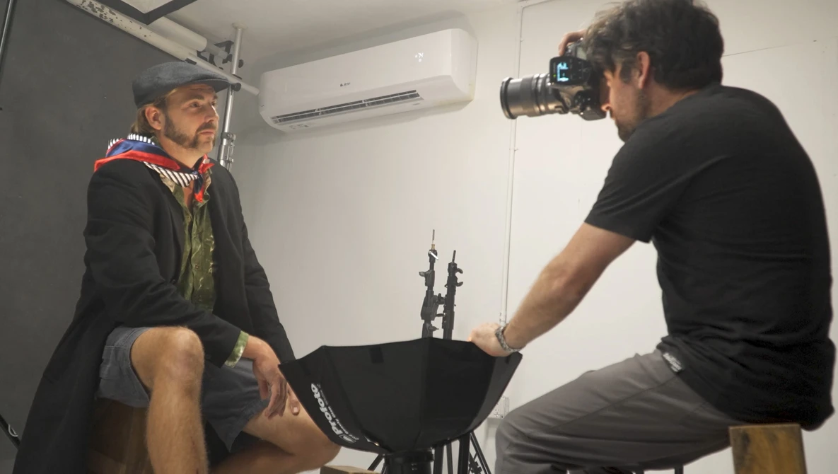 Photographer with camera on tripod conducting a portrait shoot with subject wearing cap and layered clothing.