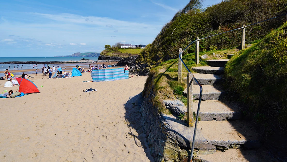 Wooden steps with rope railings descending a grassy cliff to a crowded beach with swimmers and colorful umbrellas.
