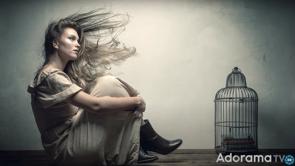 Woman sitting against a wall beside an empty birdcage, hair flowing, contemplative pose.
