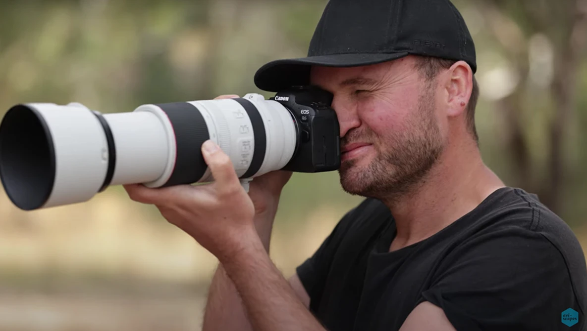 Photographer holding a telephoto lens with white barrel, looking through viewfinder in outdoor setting.