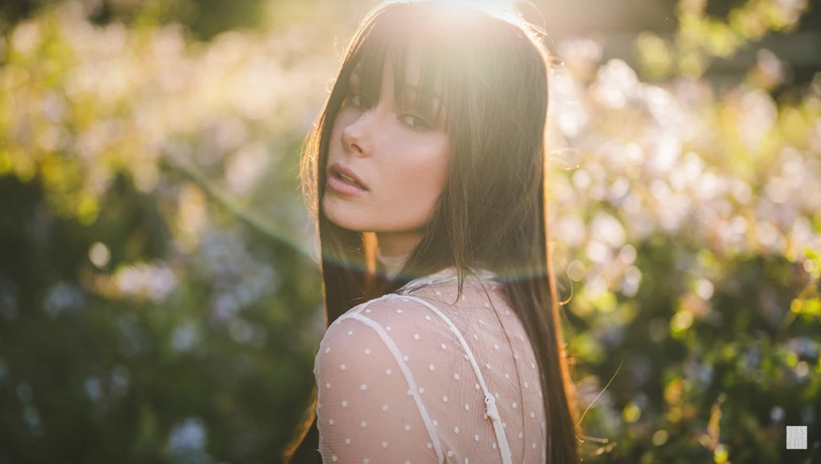Young woman with long dark hair and bangs looking over her shoulder in a sunlit wildflower field.