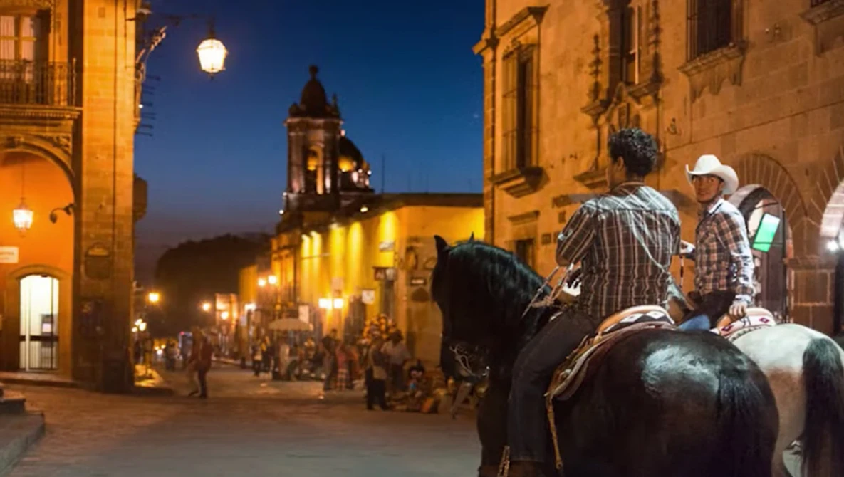 Two horseback riders in cowboy hats on a colonial town plaza at dusk, with illuminated historic buildings and a church tower in the background.