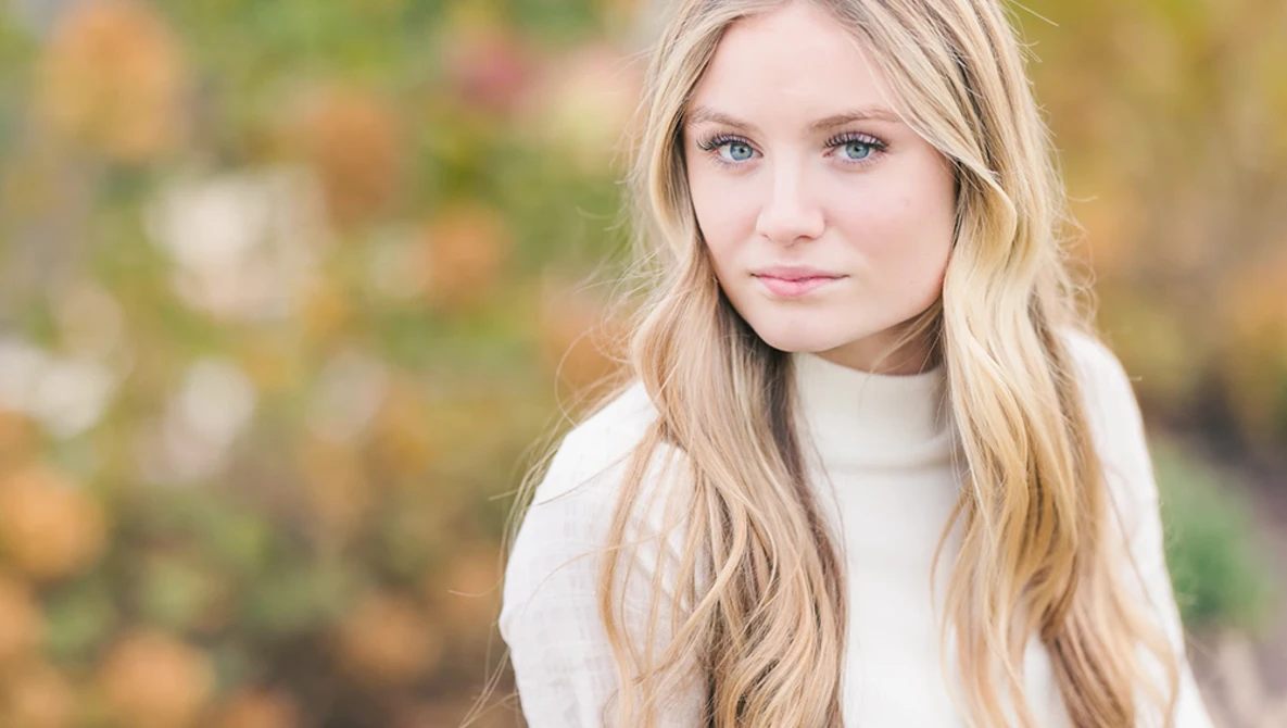 Young woman with long blonde hair wearing a white turtleneck, photographed outdoors with a blurred floral background.