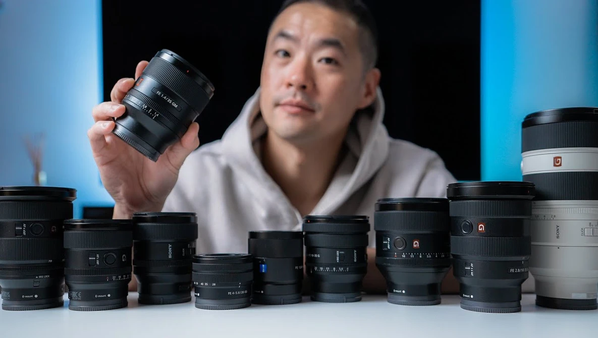 Photographer holding a camera lens above a collection of various lenses arranged on a white surface.
