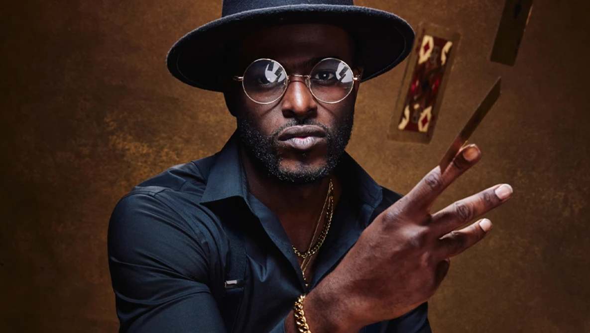 Man wearing black hat and round glasses gestures expressively against warm brown backdrop.