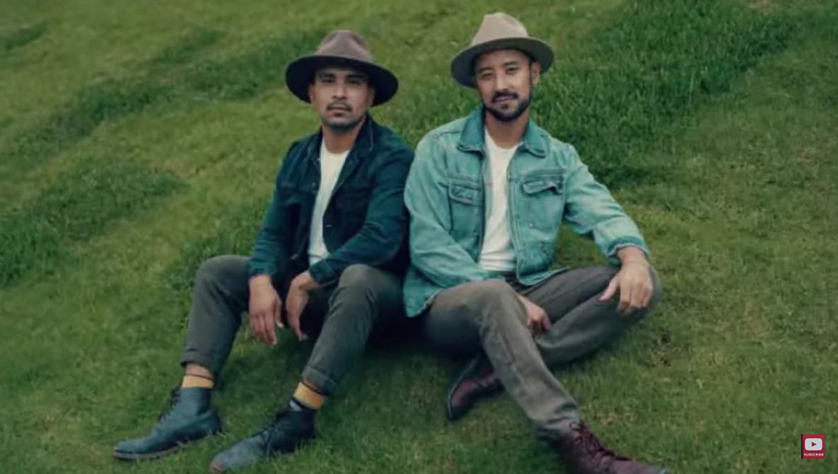 Two men sitting on grass wearing hats and casual shirts, looking at camera.
