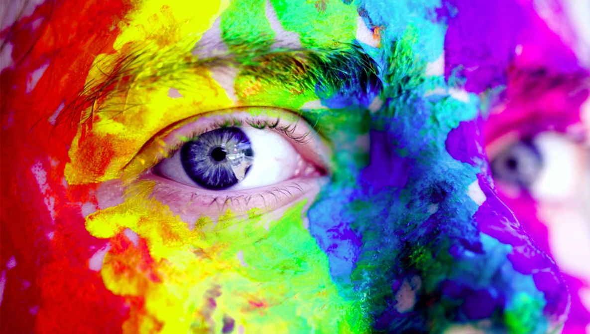 Close-up of a human eye surrounded by vibrant rainbow paint splashes and powder in red, yellow, green, blue, and purple hues.