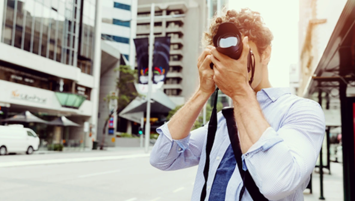 Photographer holding vintage camera up to face on urban street with tall buildings in background.