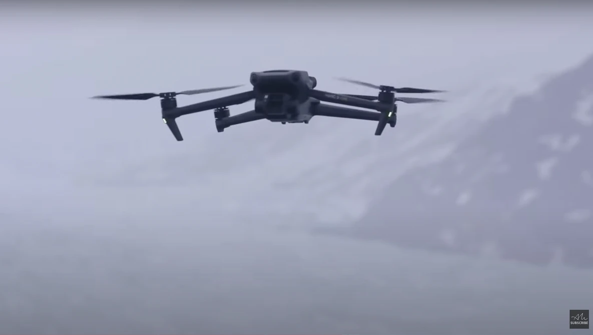 Quadcopter drone flying against an overcast sky with gray clouds in the background.