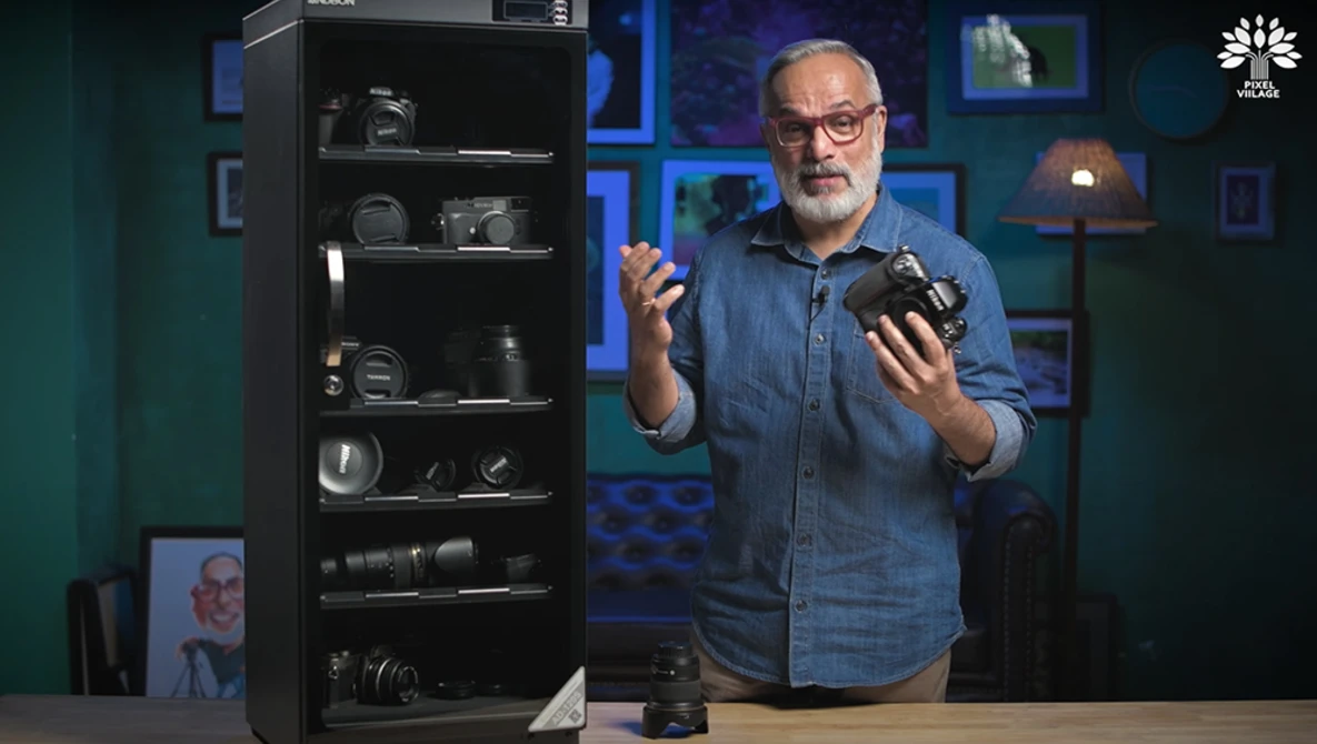 Man in denim shirt holding camera equipment while speaking in studio setting with gear display cabinet.