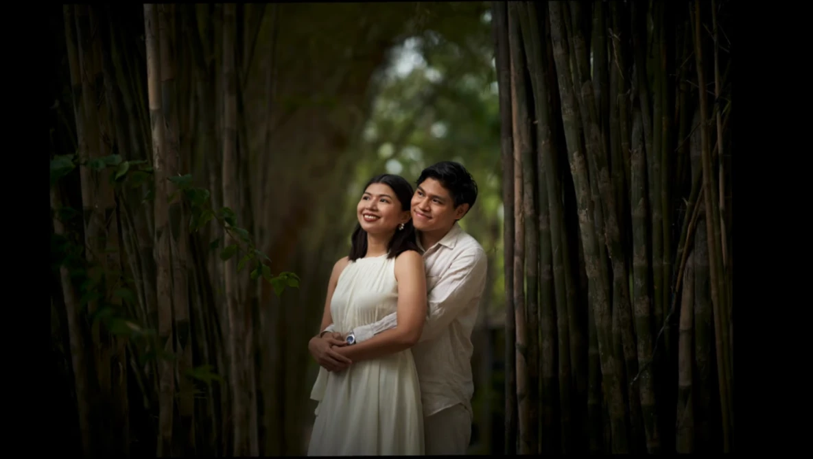 Couple embracing in a natural tunnel formed by tall trees with soft, diffused light filtering through foliage.