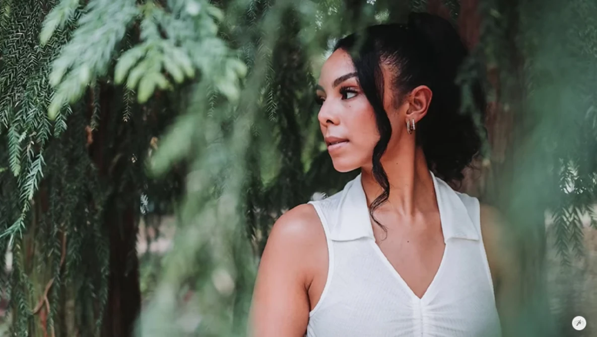 Woman in white sleeveless top among hanging green foliage, looking off-camera with natural side lighting.