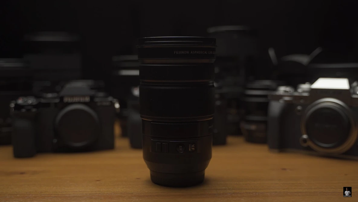 A telephoto lens in sharp focus sits centered on a wooden surface, with camera bodies blurred in the background.