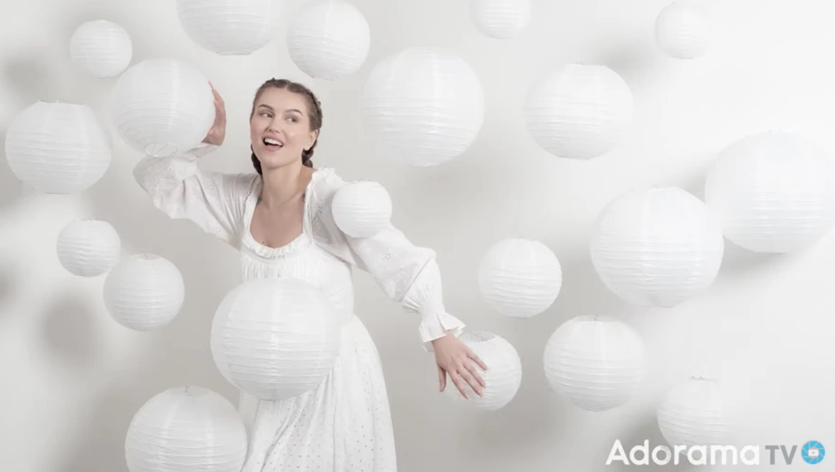 Pregnant woman in white dress posing among floating white paper lanterns against a white backdrop.