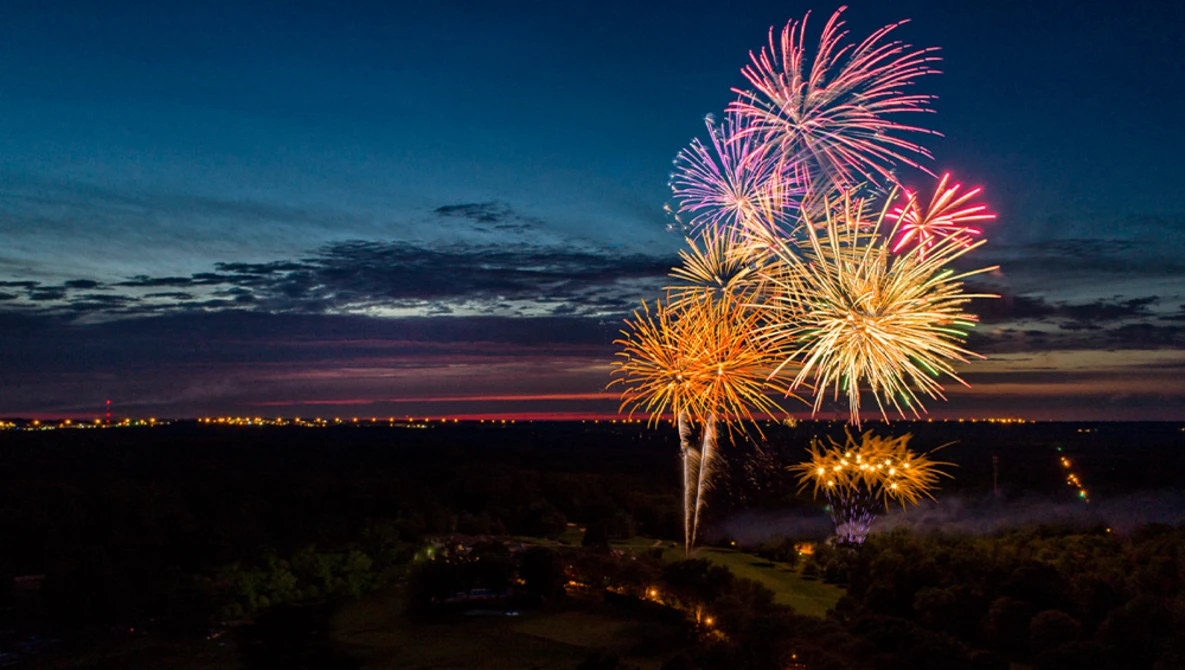 Colorful fireworks burst against a twilight sky above a darkened landscape with scattered lights.
