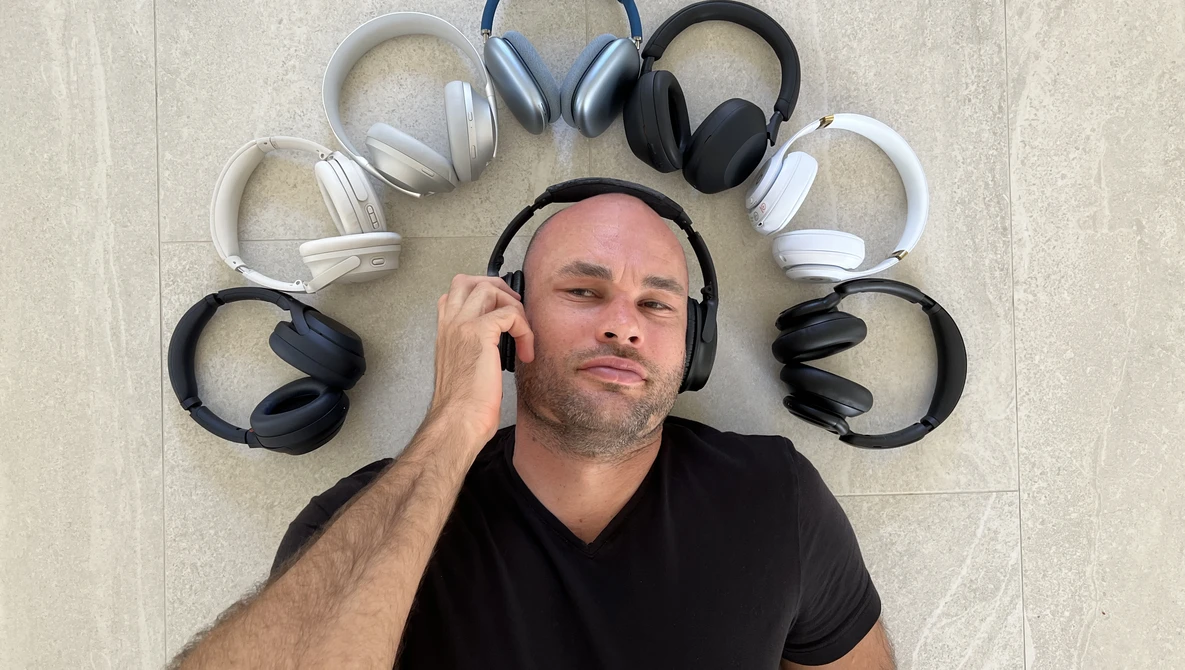 Man wearing black headphones surrounded by nine other headphones arranged in a circle on a light tile floor.