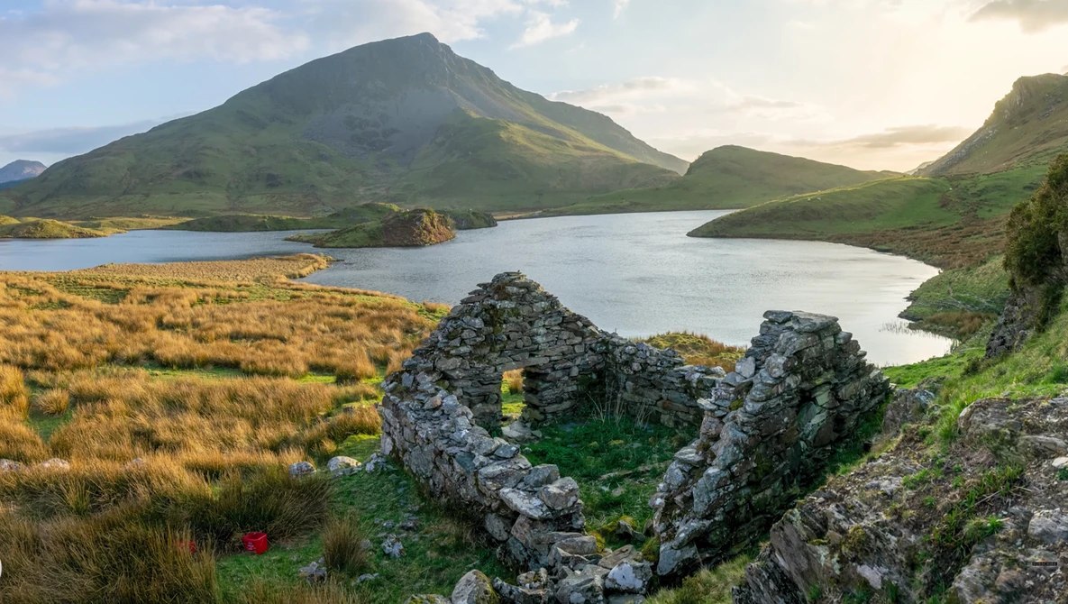 Rugged mountain landscape with cone-shaped peak, glacial lakes, and ancient stone ruins in foreground.