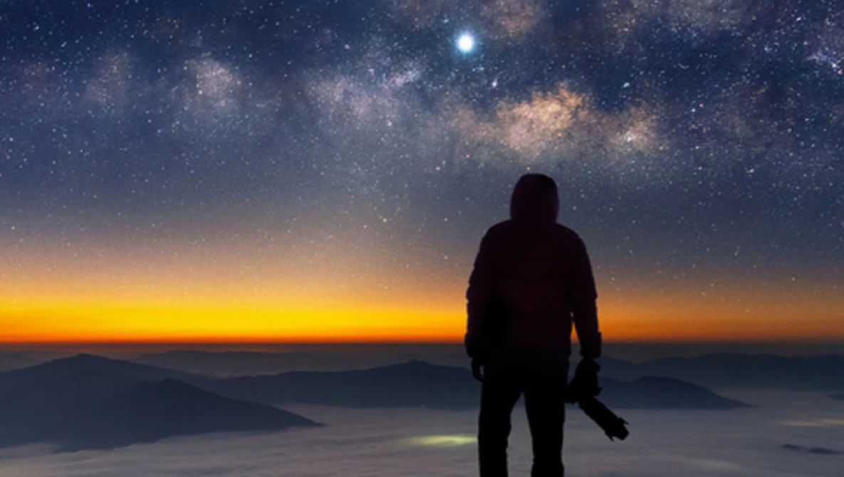 Silhouetted figure standing on a mountain ridge overlooking a sea of clouds beneath a star-filled night sky and glowing horizon.