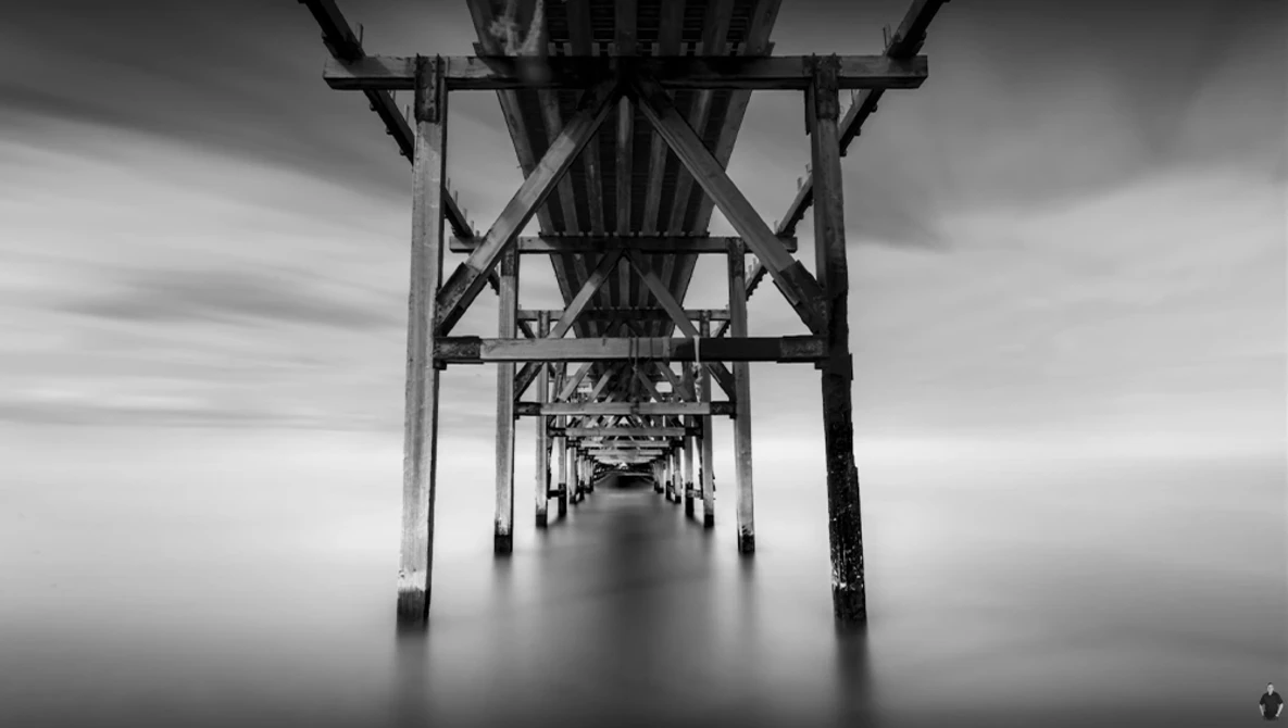 Symmetrical black and white photograph taken underneath a pier, showing repeating wooden support structures receding into a misty distance.