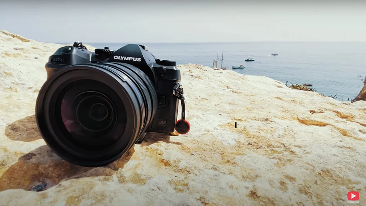 Olympus camera with large lens resting on sandy beach terrain overlooking anchored boats in calm water.