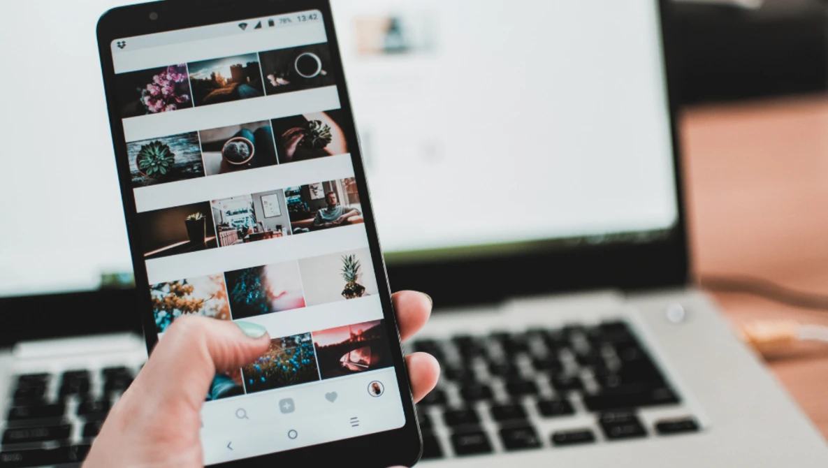 Smartphone displaying a grid of colorful photographs in a gallery app, held in hand above a laptop.