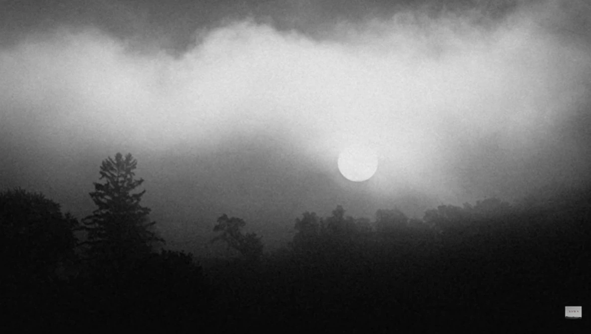 Sun obscured by heavy storm clouds above a dark forested landscape at dusk or dawn.