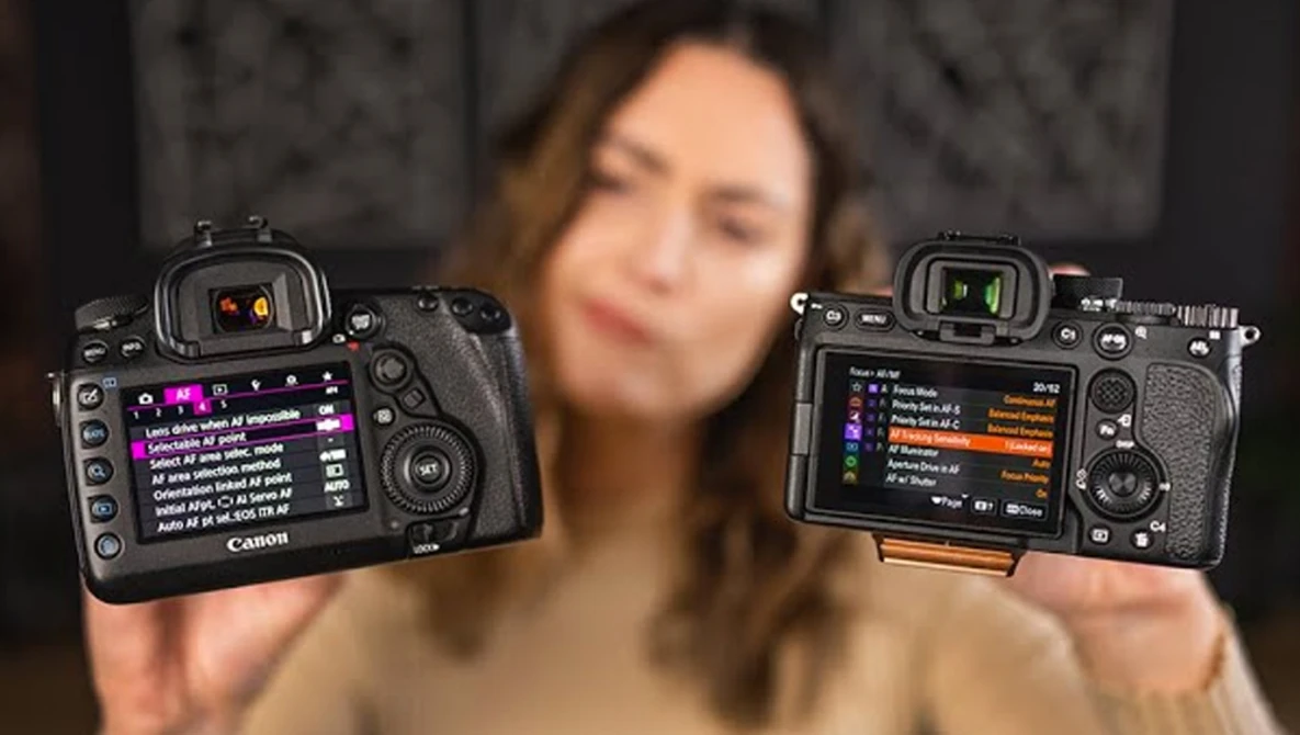 Two Canon mirrorless cameras held up side by side, displaying rear LCD screens with menu interfaces and settings visible.