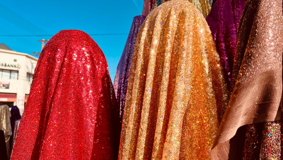 Vibrant sequined fabrics in red, gold, and burgundy draped and displayed outdoors in bright sunlight.