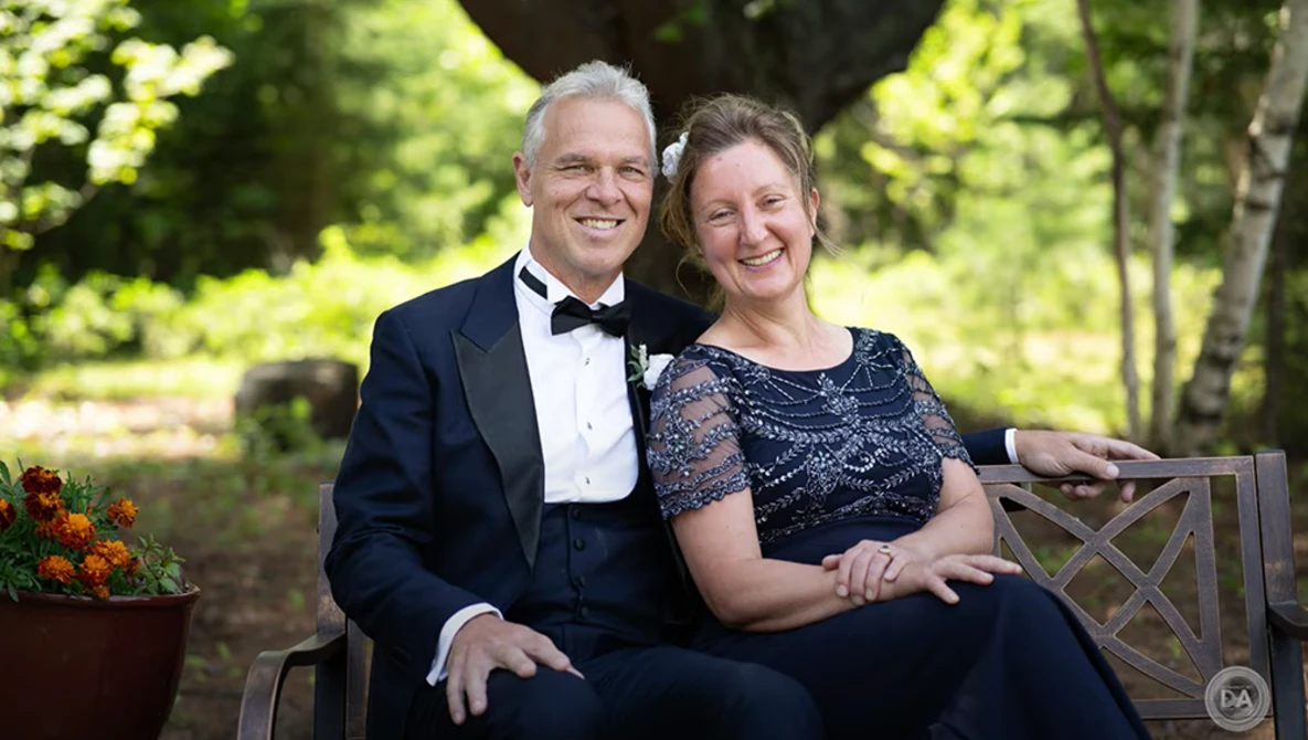 Couple in formal attire sitting on a wooden bench outdoors, smiling at camera.