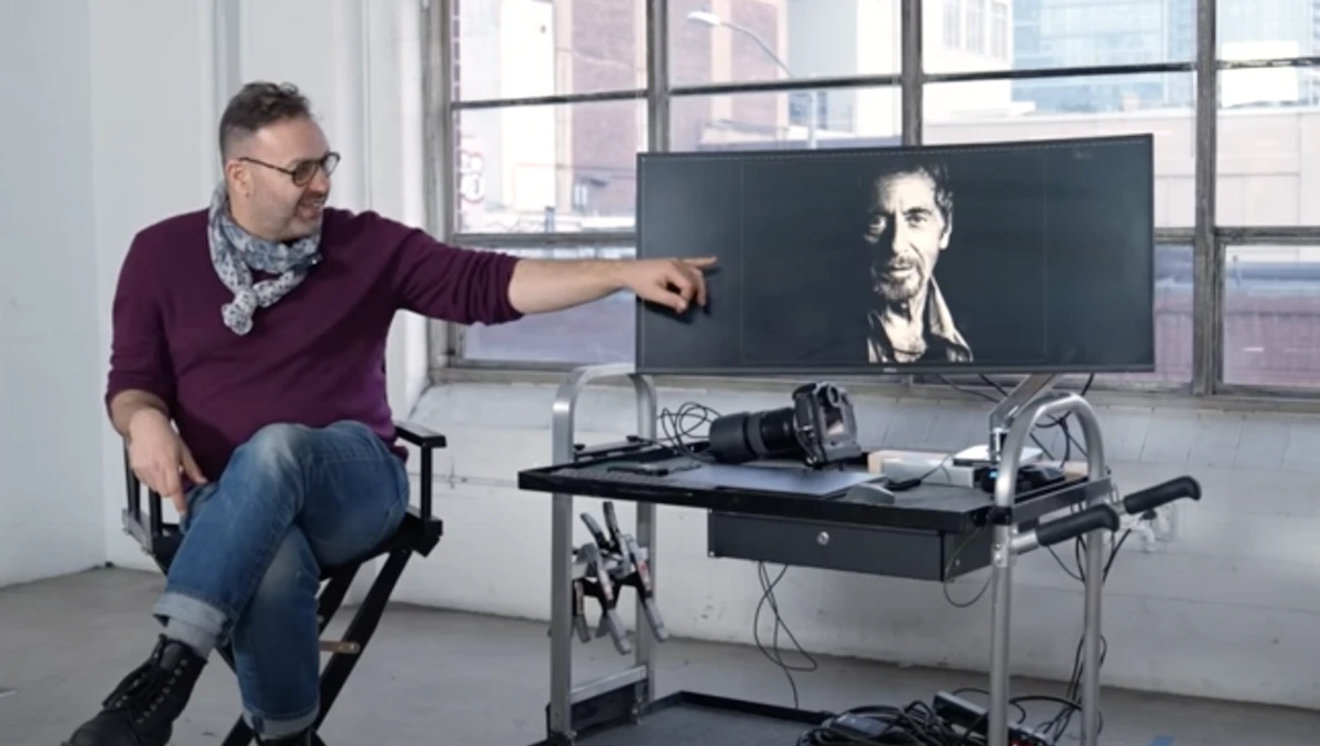 Man in burgundy sweater points at portrait displayed on wide monitor at editing workstation.