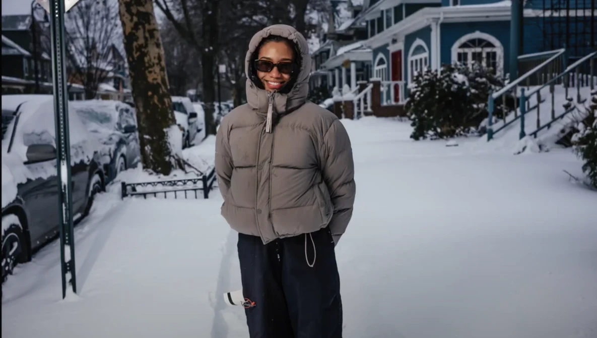 Person wearing sunglasses and a beige puffer jacket standing in a snowy urban street.