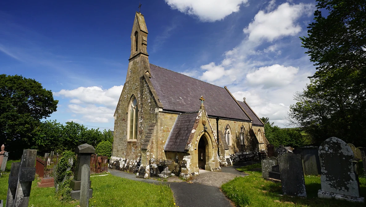 Stone chapel with steep pitched roof and tall spire surrounded by weathered gravestones in overgrown cemetery.